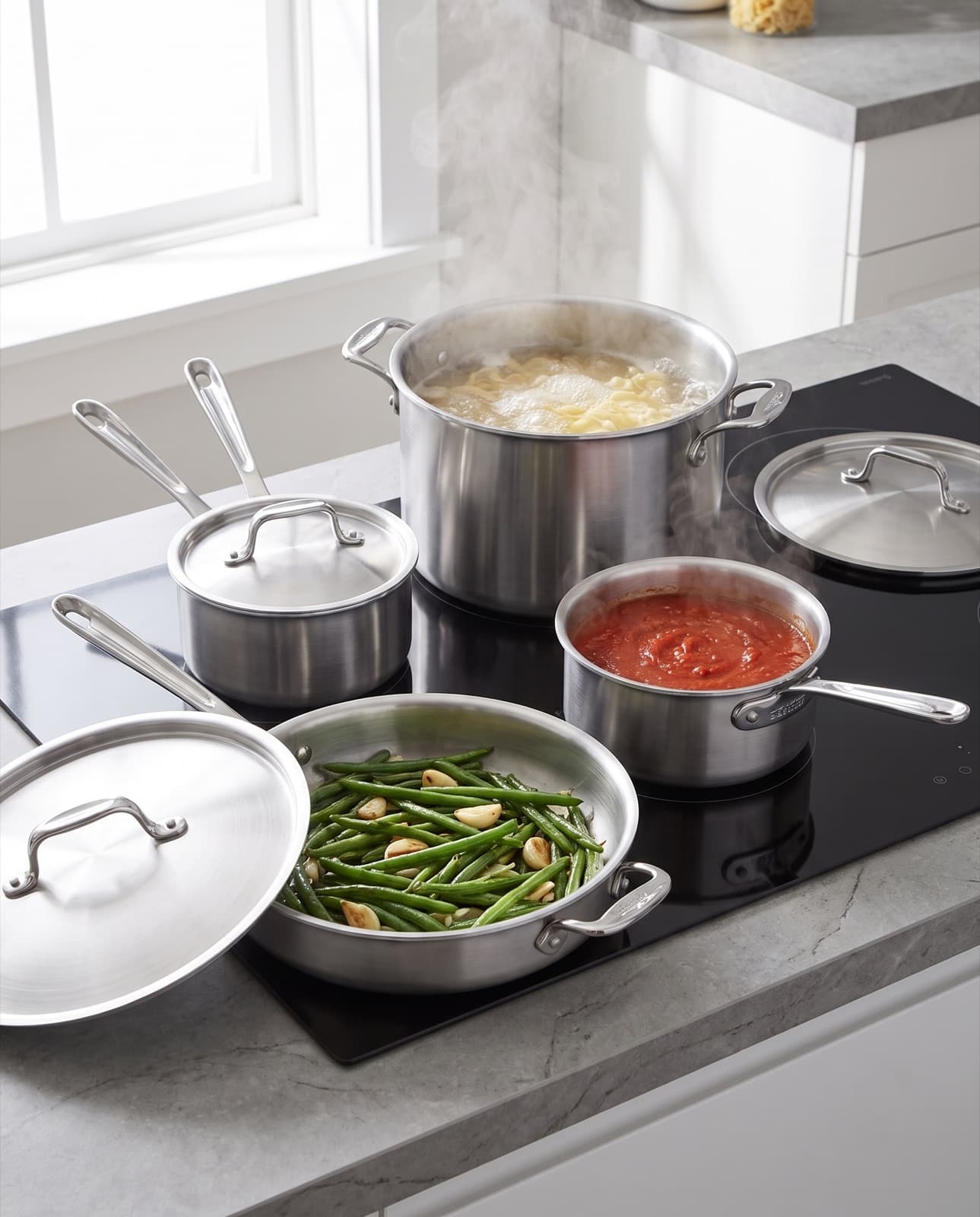 La batterie en inox sur une plaque à induction : pâtes dans le faitout fumant, sauce tomate, haricots verts à la sauteuse