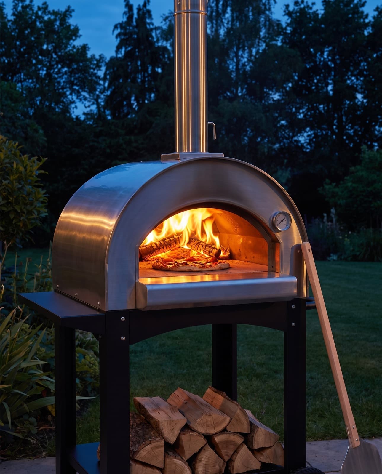 Le four à bois allumé dans un jardin à l'heure bleue, feu vif au fond, pizza sur la sole et pelle en bois appuyée