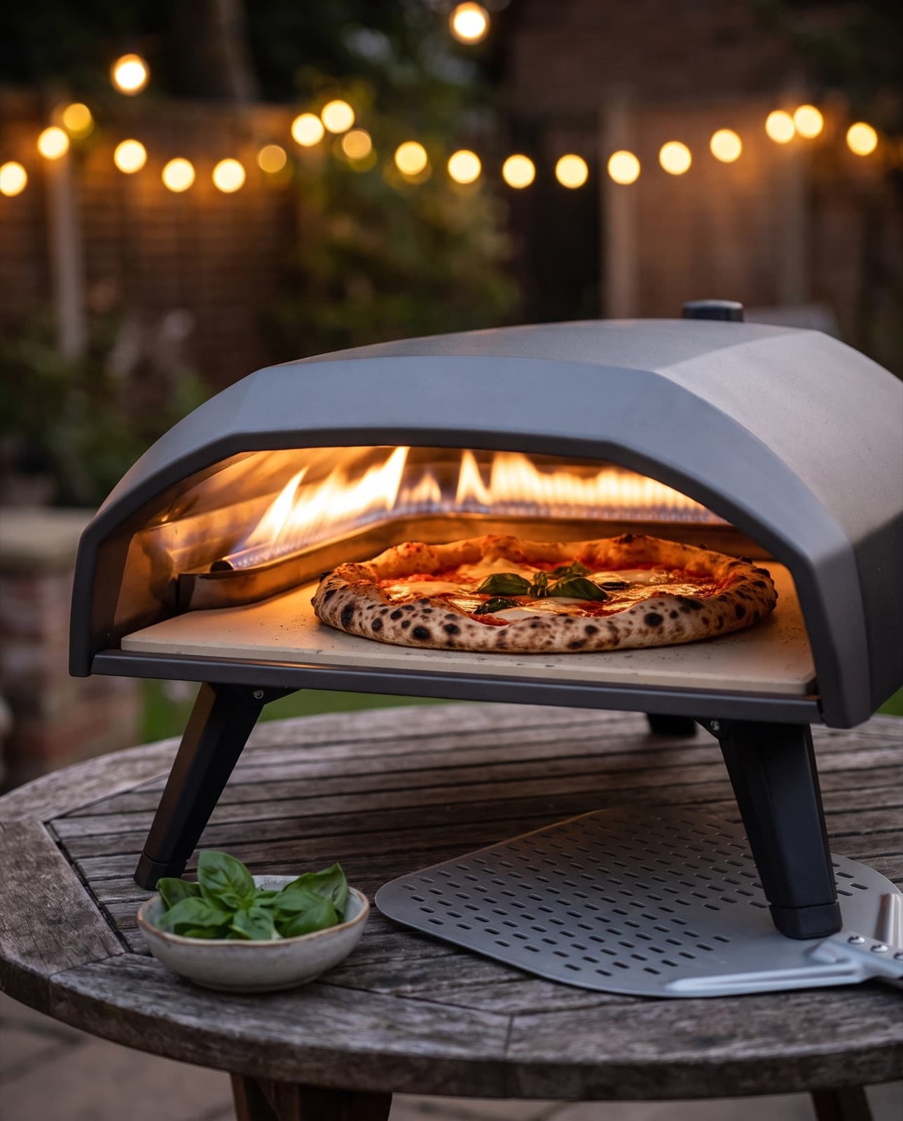 Le four allumé sur une table de jardin au crépuscule, flamme au fond et pizza napolitaine à la croûte tachetée